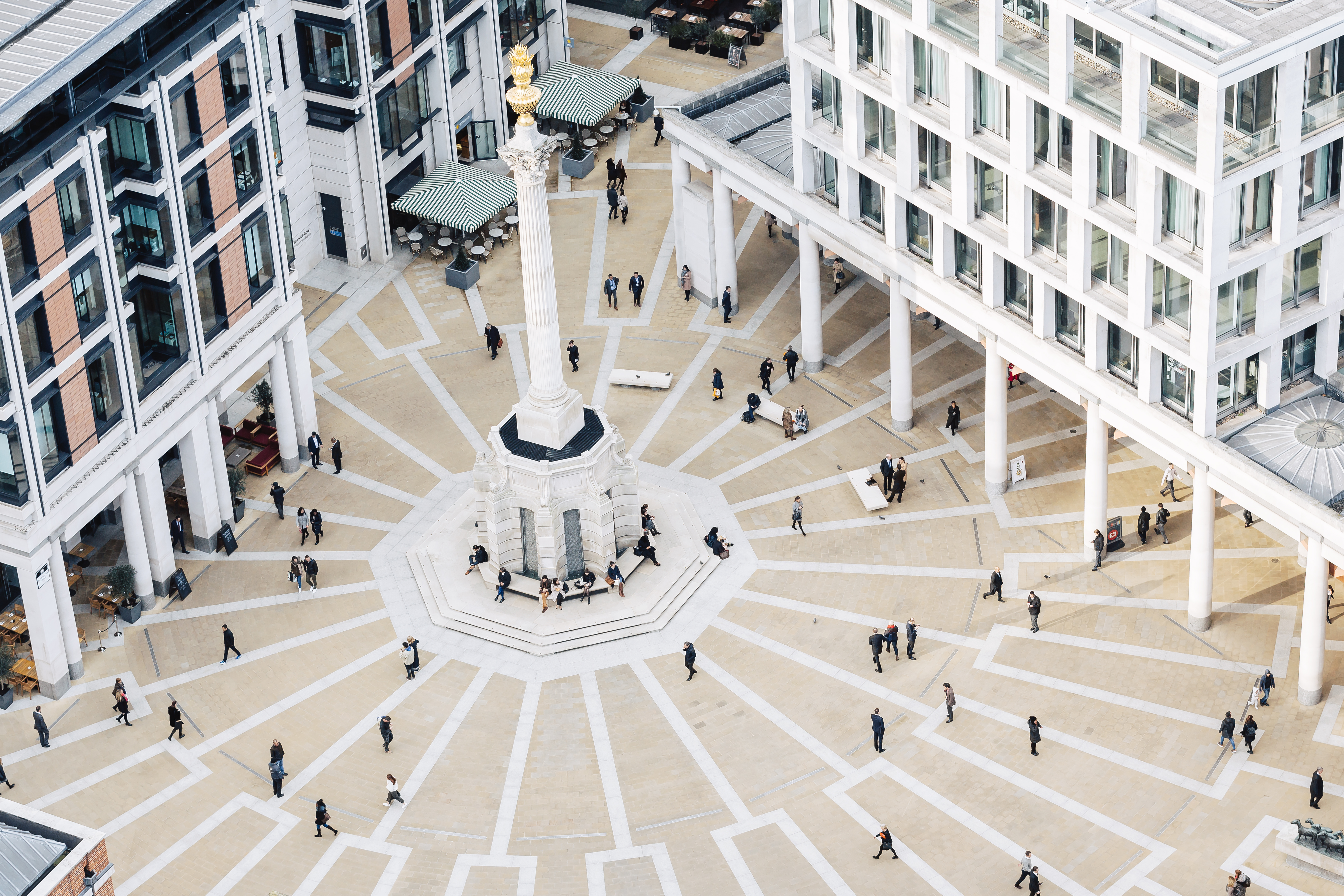 Aerial view of Paternoster Square in London, showing the central column, radial paving patterns, surrounding modern buildings, and people walking or gathering in the public space.