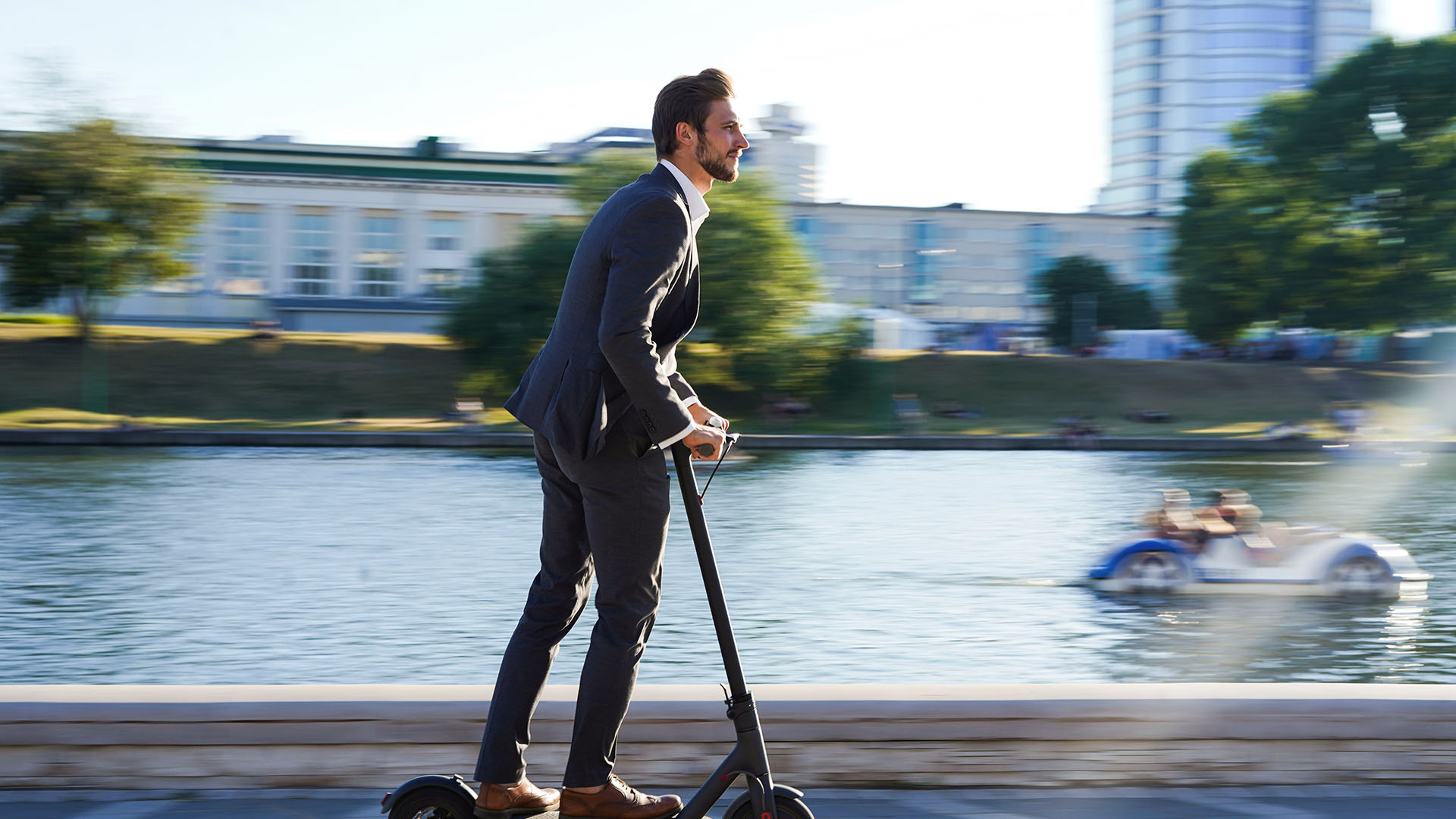 A man in a formal suit rides an electric scooter along a riverside pathway. 