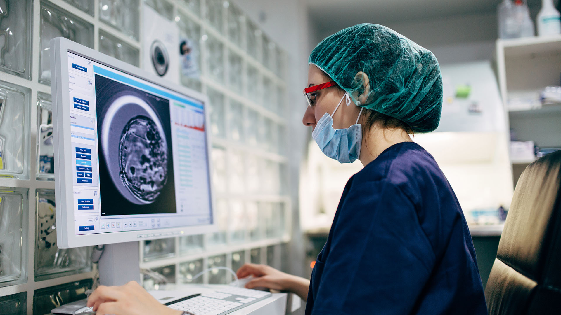 A healthcare worker wearing a surgical cap and scrubs is seated at a desk, examining detailed medical imaging on a computer screen. 