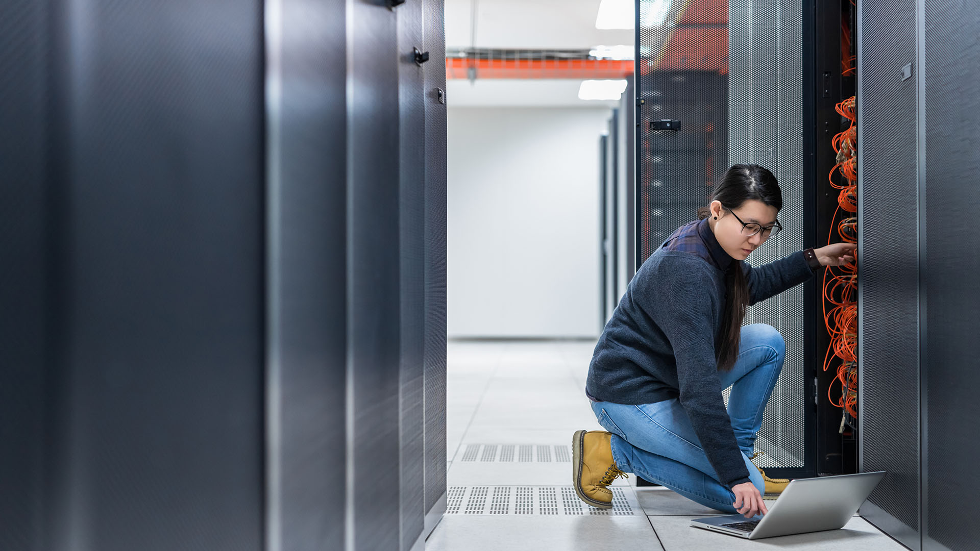 A technician is kneeling in a modern server room, working on a laptop while inspecting network cables.